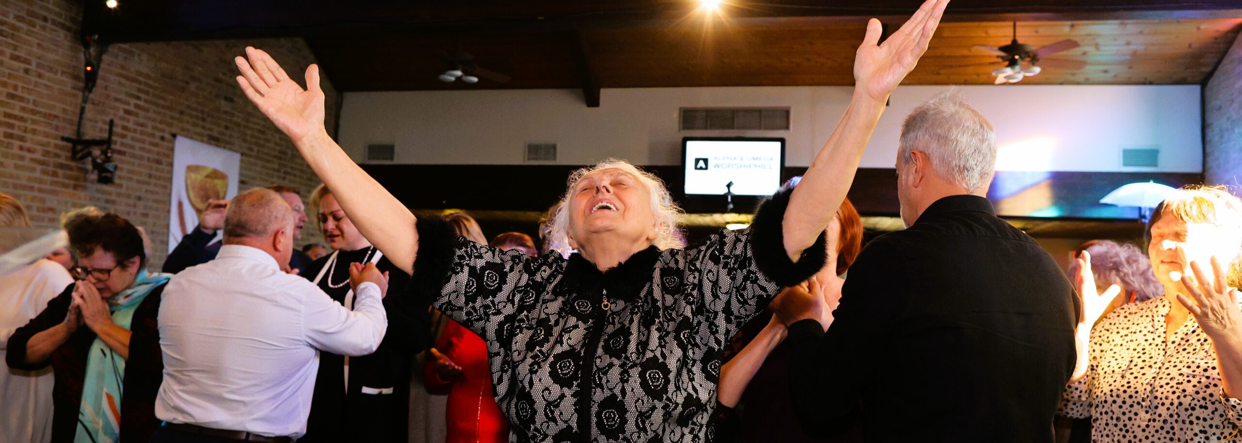 An elderly woman with arms raised and eyes closed stands joyfully among a group of people in a warmly lit indoor space, with others around her also engaged in emotional or celebratory gestures.