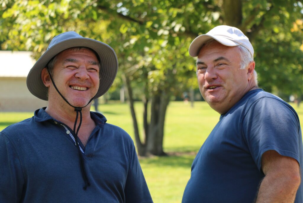 Two middle-aged men stand outdoors on a sunny day, both smiling and wearing hats. Trees and a grassy area are in the background, suggesting a park setting.