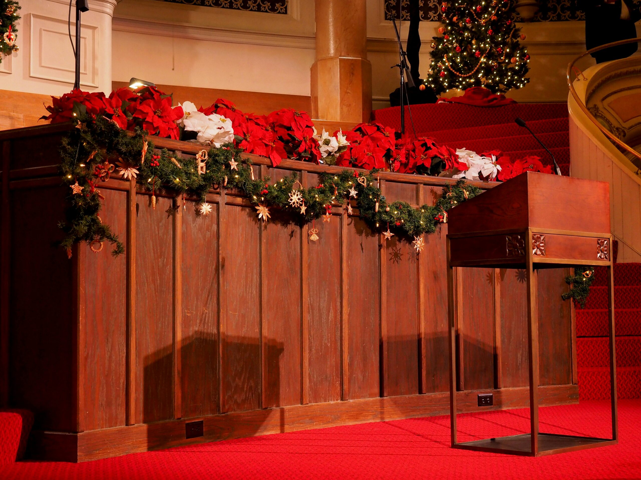 A wooden podium stands on a red-carpeted stage decorated with garlands, ornaments, and poinsettias, with a Christmas tree and festive lights in the background.