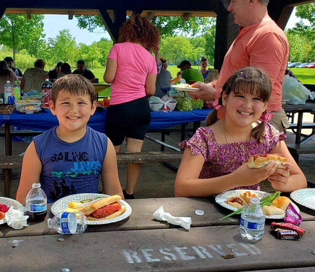 Three people—an older woman, a boy, and a girl—sit at a picnic table with plates of food, smiling at the camera. Others are gathered in the background under a park pavilion on a sunny day.