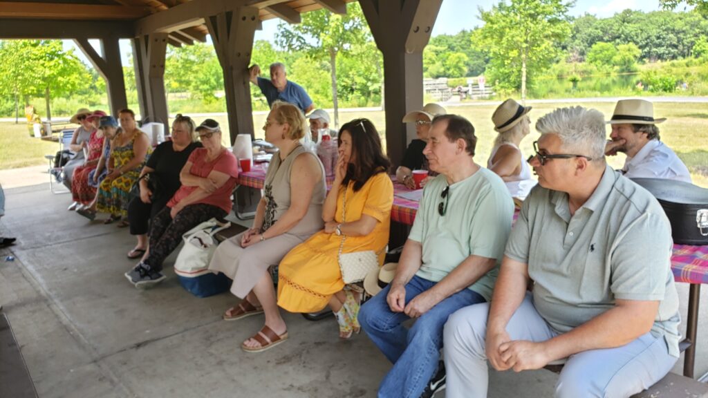 A group of people sit on benches under a pavilion in a park, attentively facing the same direction. The weather is sunny and the background features green trees and grass.