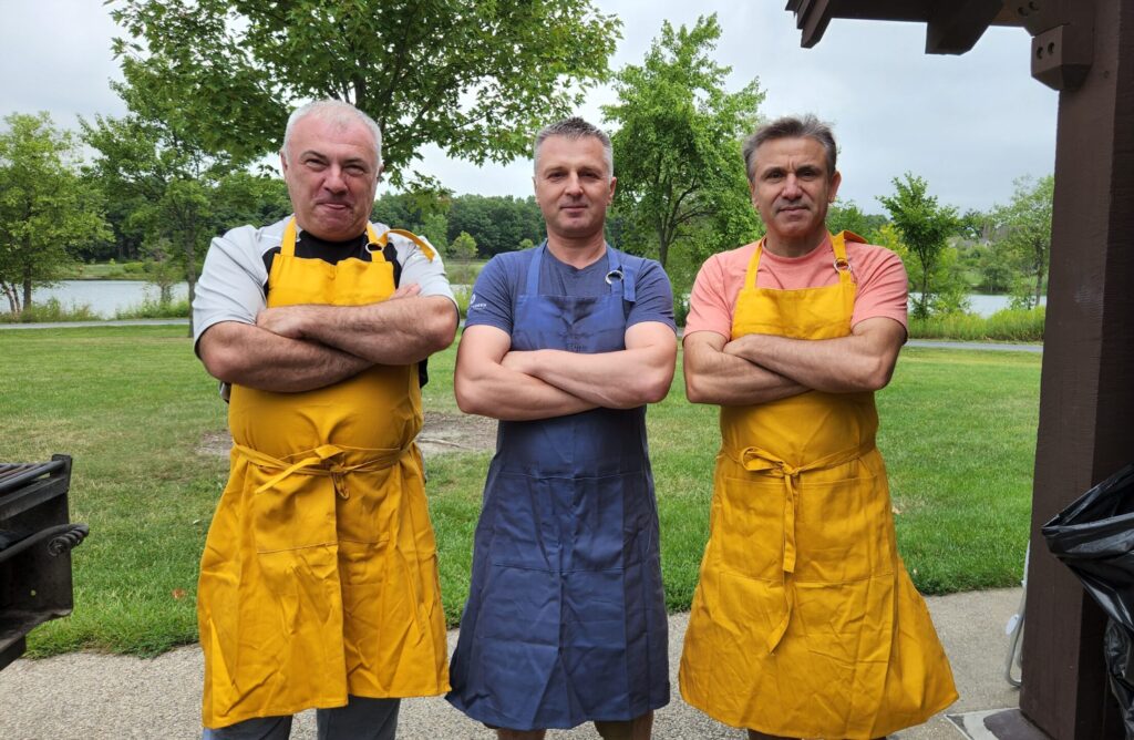 three men stand side by side outdoors, each with arms crossed. they wear yellow aprons over casual clothes and pose in front of green grass, trees, and a lake on a cloudy day.