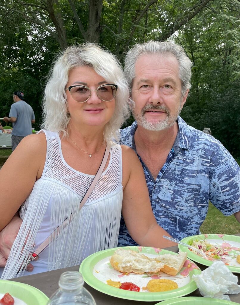 A smiling couple sits at a picnic table outdoors, with food and water bottles on green paper plates in front of them. Trees and other people are visible in the background on a sunny day.